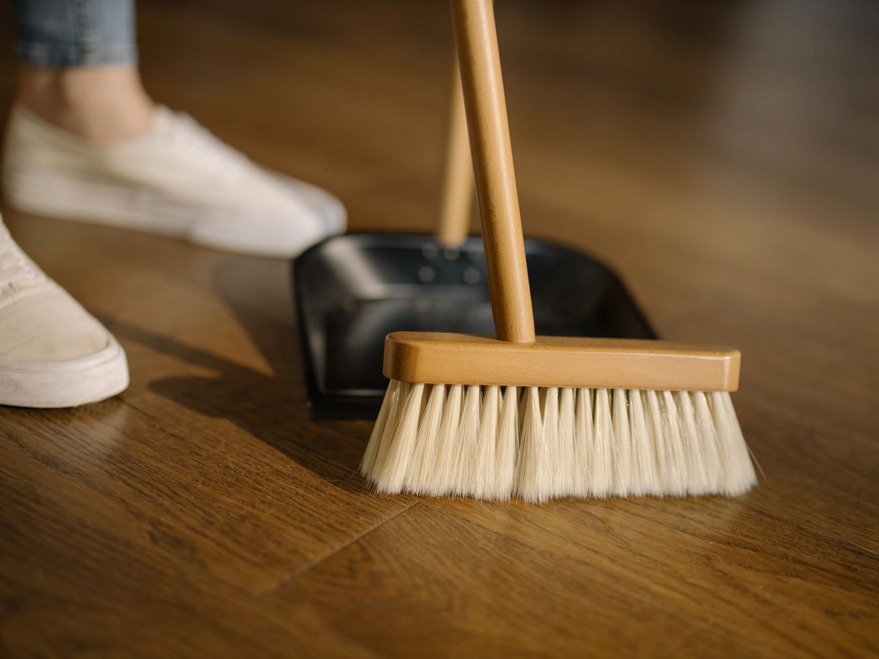 portfolio-05 Close-up of a broom and dustpan with white sneakers indoors, representing housekeeping and cleanliness.